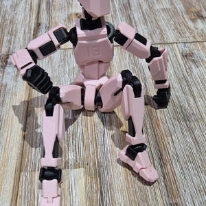 May include: A pink articulated action figure with black joints, sitting with its legs crossed. The figure has a simple, blocky design.