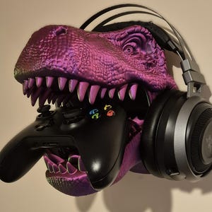 May include: A purple dinosaur head wall mount with a black gaming headset and a black video game controller in its mouth.