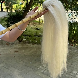 May include: A light-colored, long-haired brush with a bamboo handle. The handle is a light brown color with a natural bamboo texture. The brush is held by a person in a white shirt. The background is a mix of greenery and a paved surface.