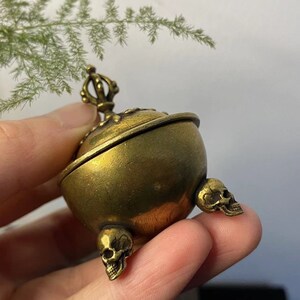 May include: A small, antique brass incense burner with a spherical body and three skull-shaped feet. The lid is ornate, and the burner is held in a hand. The overall design is detailed and gothic.