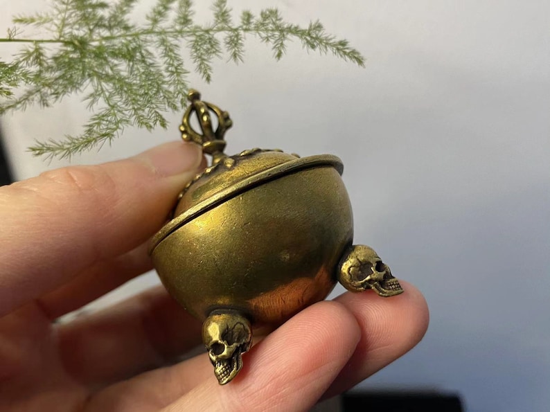 May include: A small, antique brass incense burner with a spherical body and three skull-shaped feet. The lid is ornate, and the burner is held in a hand. The overall design is detailed and gothic.
