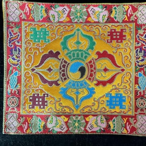 May include: A square, ornate textile with a central design featuring a yin-yang symbol. The textile has a yellow background with blue, green, and maroon accents. The border is red with floral and geometric patterns.