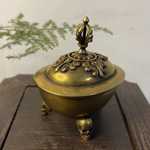 May include: A brass incense burner with a decorative lid and three skull-shaped feet. The lid features ornate detailing and a finial. The burner has a golden hue and sits on a wooden surface. A sprig of green foliage is in the background.