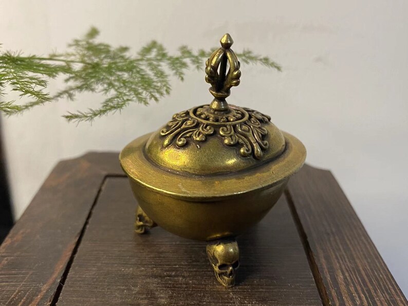May include: A brass incense burner with a decorative lid and three skull-shaped feet. The lid features ornate detailing and a finial. The burner has a golden hue and sits on a wooden surface. A sprig of green foliage is in the background.