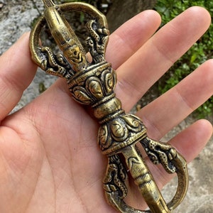 Op de afbeelding: Een gedetailleerde, antieke messing vajra, een ritueel object met ingewikkelde ontwerpen. De vajra heeft een centrale bol, sierlijke details en gebogen tanden, met een verweerde, verouderde uitstraling. Het object wordt in een hand gehouden.