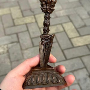 Può includere: Un pugnale rituale phurba color bronzo scuro con una testa di divinità a tre facce sulla sommità, un'impugnatura centrale con intagli complessi e una base a lama triangolare su un piedistallo decorativo, a gradini e smerlato.
