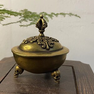 May include: A bronze-colored incense burner with a decorative lid and three skull-shaped feet. The lid is adorned with intricate patterns and a central finial. The burner sits on a wooden surface.