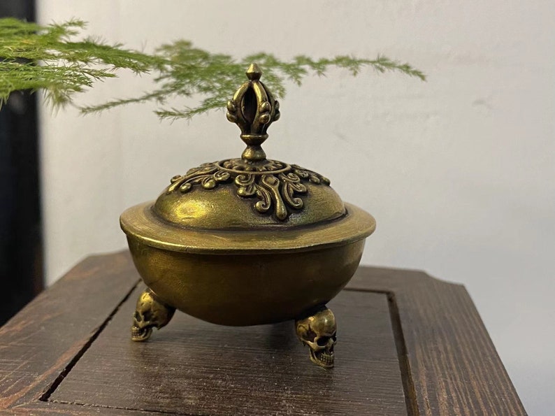 May include: A bronze-colored incense burner with a decorative lid and three skull-shaped feet. The lid is adorned with intricate patterns and a central finial. The burner sits on a wooden surface.
