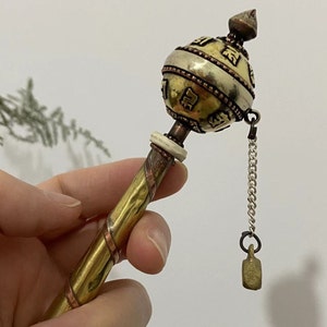 Puede incluir: Una rueda de oración de mano con un mango de latón y cobre, un cuerpo cilíndrico con símbolos grabados y una pequeña campana sujeta por una cadena. La rueda de oración tiene una combinación de colores dorados, plateados y cobrizos.