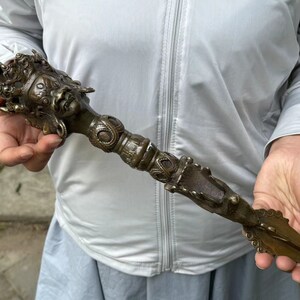 Puede incluir: Una daga ritual phurba de color bronce que presenta una compleja cabeza de deidad de tres caras en la parte superior, bandas decorativas ornamentadas a lo largo del mango cilíndrico y una hoja triangular afilada en la base con metal detallado.