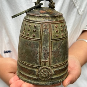 Può includere: Un'antica campana di bronzo con manico e battaglio. La campana ha una forma arrotondata con rivetti decorativi e un motivo floreale alla base. La campana è tenuta tra le mani di una persona.