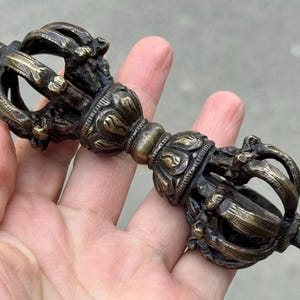 Peut inclure: Vajra en bronze orné, un objet rituel aux détails complexes. Le vajra présente une tige centrale avec des éléments décoratifs et des pointes symétriques à chaque extrémité. L'objet est tenu dans une main.
