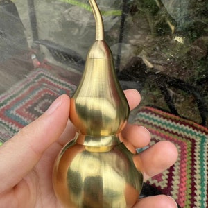 May include: A gold-coloured gourd-shaped container with a curved spout. The container has a smooth, polished surface and a bulbous bottom. The gourd is held in a hand, with a colourful patterned background visible.