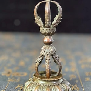 Könnte beinhalten: Ein bronzefarbener, verzierter buddhistischer Vajra, ein Ritualobjekt. Der Vajra hat ein zentrales zahnförmiges Element, einen dekorativen Griff und eine lotusförmige Basis. Das Metall hat ein verwittertes Aussehen, das auf Alter oder einen Vintage-Stil hindeutet.