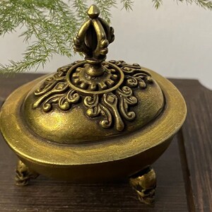 May include: A gold-colored decorative incense burner with a detailed lid and skull-shaped feet. The lid features ornate patterns and a crown-like finial. The burner is set against a dark wooden surface.