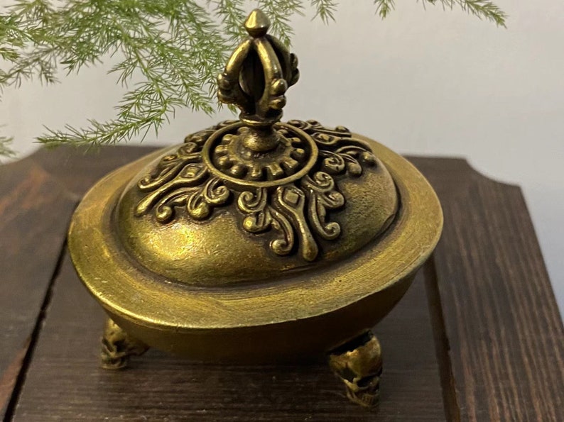 May include: A gold-colored decorative incense burner with a detailed lid and skull-shaped feet. The lid features ornate patterns and a crown-like finial. The burner is set against a dark wooden surface.