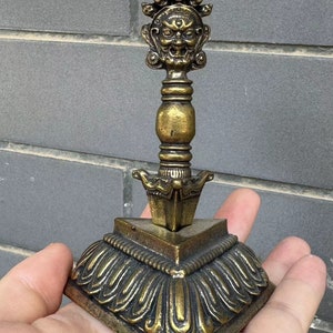 Puede incluir: Un objeto ritual de bronce ornamentado con un diseño detallado. La parte superior presenta una cara feroz y pequeñas calaveras, mientras que la base tiene un patrón en capas en forma de pétalos. El objeto se sostiene en una mano, mostrando su tamaño.