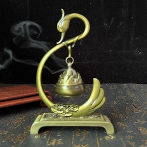 Peut inclure: Un brûleur d'encens en laiton en forme de cygne. Le brûleur a un porte-encens en forme de cloche suspendu au cou du cygne. La base est rectangulaire avec un motif floral. De la fumée s'élève en arrière-plan.