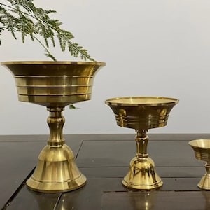 May include: Three brass-colored decorative bowls of varying sizes. The bowls have a flared rim and a pedestal base. The largest bowl is approximately 6 inches tall, the medium bowl is 4 inches tall, and the smallest is 2 inches tall. The bowls are displayed on a dark wooden surface.