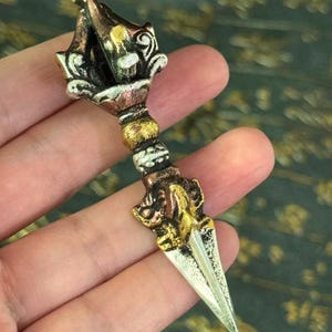 Pode incluir: Uma adaga ritual phurba de metal em tons múltiplos com uma lâmina pontiaguda, um punho central com faixas texturizadas e uma seção superior ornamentada. O objeto exibe uma mistura de acabamentos em cobre, latão e cor prateada.