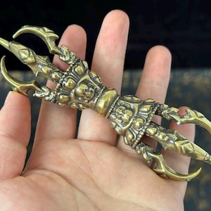 Peut inclure: Un vajra en laiton détaillé, un objet rituel avec une poignée centrale et des griffes s'étendant de chaque extrémité. Le vajra est tenu dans une main, mettant en valeur son design complexe et sa couleur dorée. Le fond est sombre.