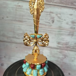 May include: A gold-colored decorative object with a detailed design. The object features turquoise and red stones, with an ornate top. The base is round and the overall design is intricate and eye-catching.