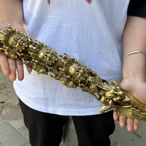 May include: A detailed, golden ritual dagger with intricate carvings of faces and figures. The dagger is held in a person's hands, showcasing its ornate design and sharp blade. The handle features a bulbous top.