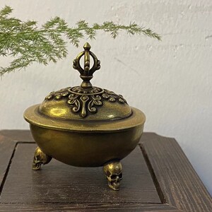 May include: A bronze-colored incense burner with a decorative lid and three skull-shaped feet. The lid is adorned with intricate patterns. The burner sits on a dark wooden surface, with green foliage in the background.