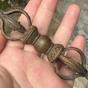 May include: A bronze-colored Vajra, a ritual object with a central sphere and stylized prongs. The intricate details include floral and geometric patterns. The object is held in a hand, showcasing its size and craftsmanship.