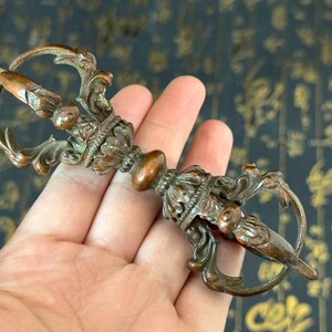 May include: An ornate bronze ritual dagger with detailed floral and scrollwork. The double-sided blade has a pointed tip. The handle has a bulbous centre and ring-shaped ends. A decorative religious item.