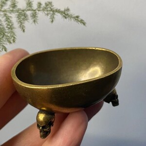 May include: A small, antique-style brass bowl with three skull-shaped feet. The bowl has an oval shape and a smooth, polished surface. The skulls are detailed and add a unique, gothic touch to the piece. The bowl is held in a hand.