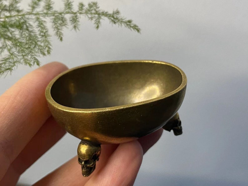 May include: A small, antique-style brass bowl with three skull-shaped feet. The bowl has an oval shape and a smooth, polished surface. The skulls are detailed and add a unique, gothic touch to the piece. The bowl is held in a hand.