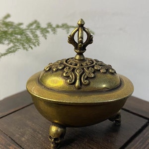 May include: A brass incense burner with a decorative lid and skull-shaped feet. The burner has an ornate design on the lid and a handle on top. The overall color is a warm, antique gold.
