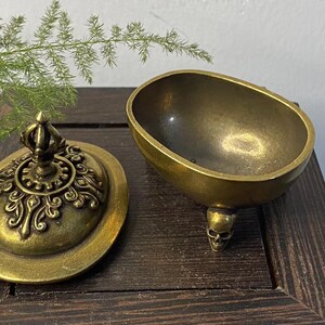 May include: A vintage brass incense burner with a skull-shaped foot. The lid is ornate with a crown-like finial and decorative patterns. The open bowl shape is oval. The burner sits on a dark wooden surface.
