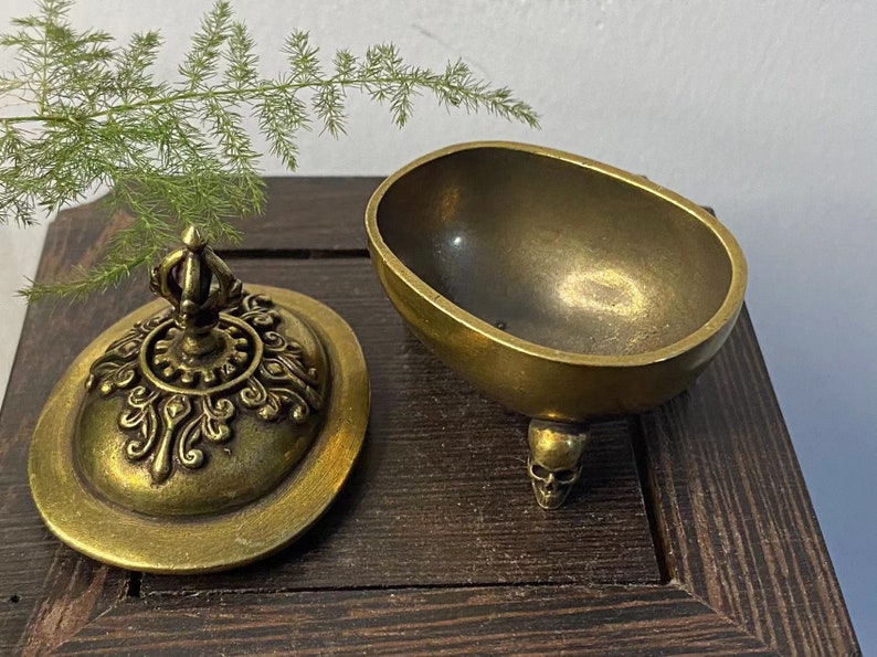 May include: A vintage brass incense burner with a skull-shaped foot. The lid is ornate with a crown-like finial and decorative patterns. The open bowl shape is oval. The burner sits on a dark wooden surface.