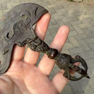 Pode incluir: Faca ritual de cor bronze com intrincadas gravuras de dragões e flores. A pega apresenta padrões detalhados e um centro bulboso. A lâmina tem uma forma curva em forma de crescente. Um item decorativo único.