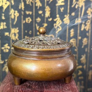 May include: A round bronze incense burner with three short legs and a removable lid. The lid features an intricate openwork floral pattern and a geometric border. A small spherical knob sits at the center of the lid, and thin wisps of smoke rise.