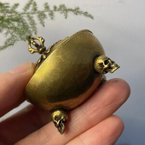 May include: A small, antique brass-colored container with skull accents. The container has a rounded shape with a decorative handle and three skull-shaped protrusions. The brass has a slightly aged appearance.