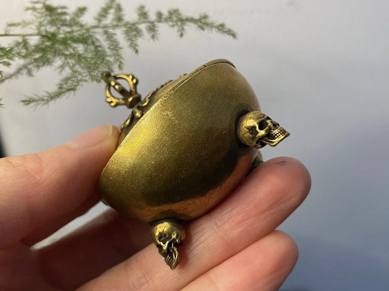 May include: A small, antique brass-colored container with skull accents. The container has a rounded shape with a decorative handle and three skull-shaped protrusions. The brass has a slightly aged appearance.