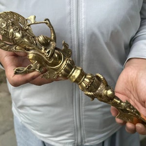 May include: A detailed brass vajra, a ritual object, with intricate carvings of deities and a dragon-like creature. The handle has a detailed design, and the pointed end is visible.