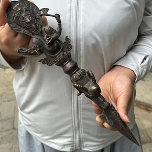 Pode incluir: Uma adaga ritual de cor bronze com entalhes intrincados. A pega apresenta uma cabeça de dragão detalhada e outras figuras ornamentadas. A lâmina é pontiaguda, sugerindo um propósito cerimonial ou decorativo.