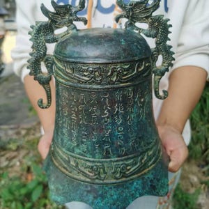 May include: An antique bronze bell with a dark patina. The bell features intricate dragon designs on the top and sides, with a curved handle. The surface is covered in engraved characters. The bell is held in front of a blurred background.
