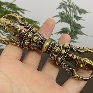 May include: A detailed brass vajra, a ritual object with a central handle and claw-like prongs. The handle features carved faces and intricate patterns. The object is held in a hand.