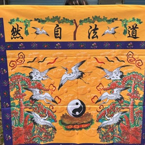May include: A decorative textile with a yellow background, featuring a central Yin and Yang symbol surrounded by cranes and stylized trees. The design includes Chinese characters and a border of purple and blue.