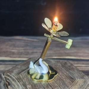 May include: A lit brass oil lamp with a flower-shaped top and a green handle. The lamp has a gold base and a white cotton wick. The flame is bright and the lamp is on a wooden surface.