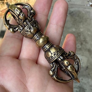 Peut inclure: Un vajra en bronze détaillé, un objet rituel, tenu dans une main. Le vajra présente des motifs complexes et une sphère centrale, avec des têtes d'animaux stylisées à chaque extrémité. Le métal a une patine chaude et vieillie.