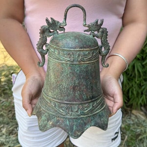 May include: An antique bronze bell with a patina finish. The bell features ornate dragon figures on the top and decorative patterns around the body. The handle is a curved bar. The bell is held in front of a blurred background.