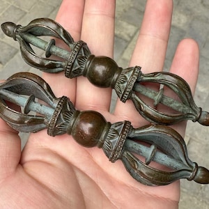 Könnte beinhalten: Zwei antike Vajra-Zepter aus Bronze, jeweils mit einem zentralen Stab und verzierten, stilisierten Enden. Die Zepter weisen dekorative Details und ein verwittertes Aussehen mit blau-grünen Patina-Akzenten auf.