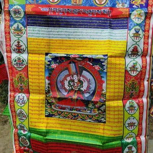 May include: A colorful Tibetan Buddhist prayer flag with intricate designs and text. The flag features a central image of a deity surrounded by detailed patterns in red, yellow, blue, and green. Decorative borders and additional religious symbols complete the design.