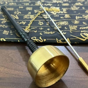 May include: A brass bell with a black handle and a gold chain, accompanied by a thin metal rod. The bell is set against a dark background with gold-colored symbols. The objects are on a wooden surface.